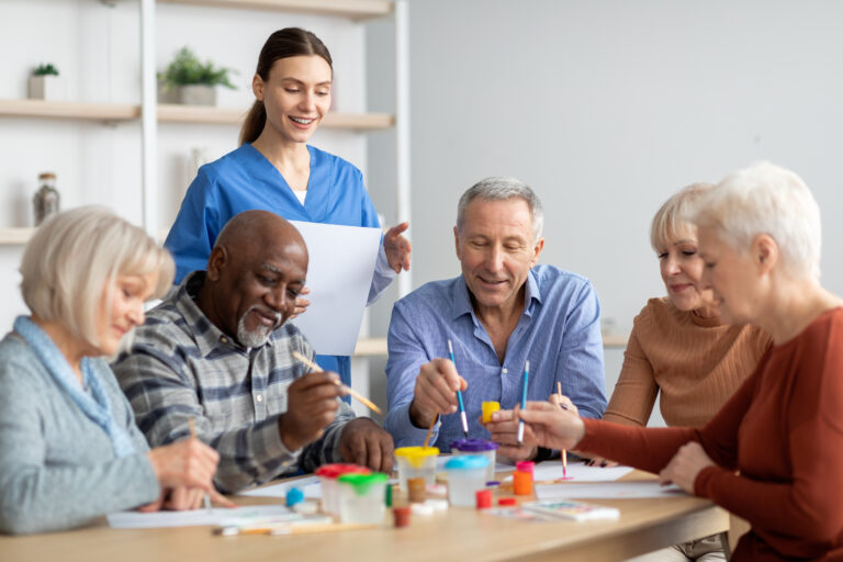 A group of elderly men and women happily engaging in arts and crafts at an adult day care center, supervised by a smiling caregiver in blue scrubs. The setting promotes social interaction and creativity for seniors. day care for elderly philadelphia