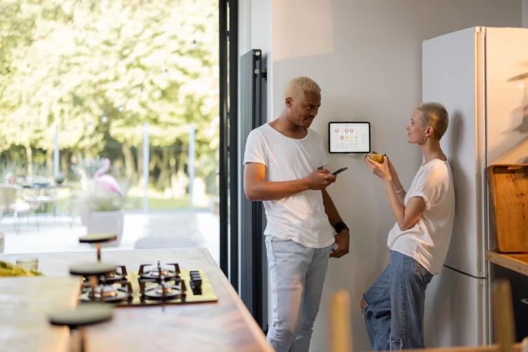 Couple managing home settings through a wall-mounted smart home control panel and smartphone.